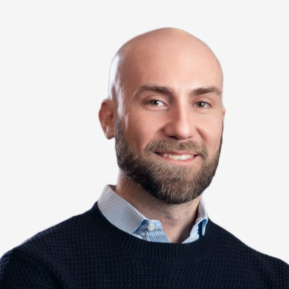 A smiling, bearded man with a bald head wearing a dark sweater over a light blue collared shirt, posing against a plain white background.