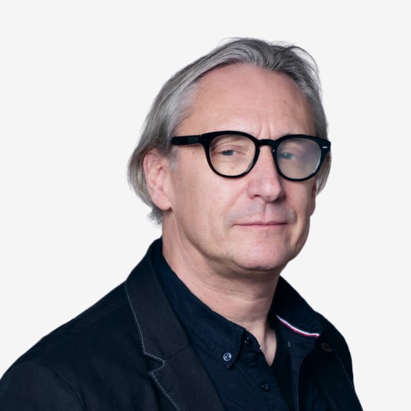 A middle-aged man with gray hair and black-framed glasses is wearing a black jacket over a dark shirt, looking slightly to the side against a plain white background.