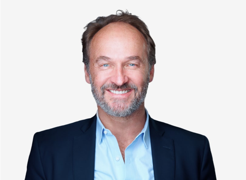 A middle-aged man with light skin, short brown hair, and a beard is smiling. He is wearing a navy blazer over a light blue shirt against a plain white background.