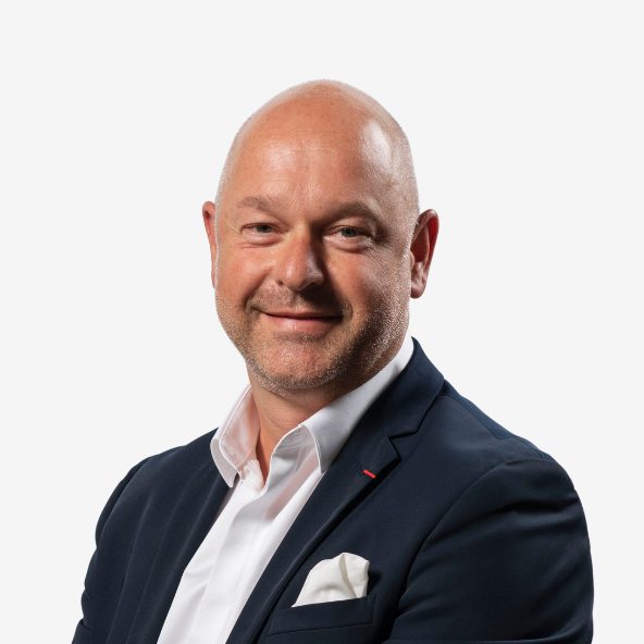 Frédéric Dalens A bald man with a trimmed beard and light complexion, wearing a dark suit jacket, white shirt, and pocket square, smiling confidently against a plain white background.